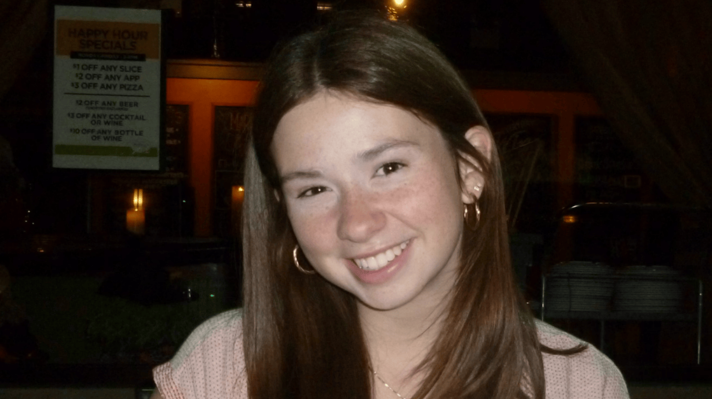 Portrait of a current Gannon Scholar with long straight brown hair and freckles, smiling, wearing hoop earrings and a light pink top, seated indoors with a softly lit restaurant setting in the background.
